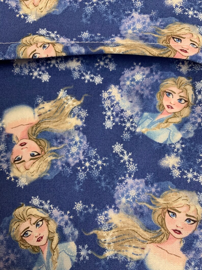 Op de afbeelding: Blauwe stof met een herhalend patroon van een cartoonfiguur met lang blond haar en een blauwe jurk. De achtergrond is een blauw en wit sneeuwvlokkenpatroon.