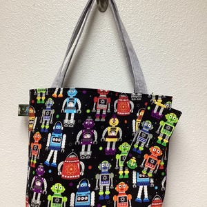 May include: A black tote bag with gray handles features a colorful pattern of robots on a black background. The robots are in various colors, including red, blue, green, yellow, and orange. The bag has a small pocket on the side.