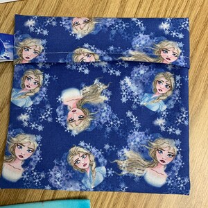 Op de afbeelding: Een blauwe stoffen tas met een wit sneeuwvlokkenpatroon en afbeeldingen van een cartoonfiguur met blond haar en blauwe ogen. De tas heeft een wit trekkoord.