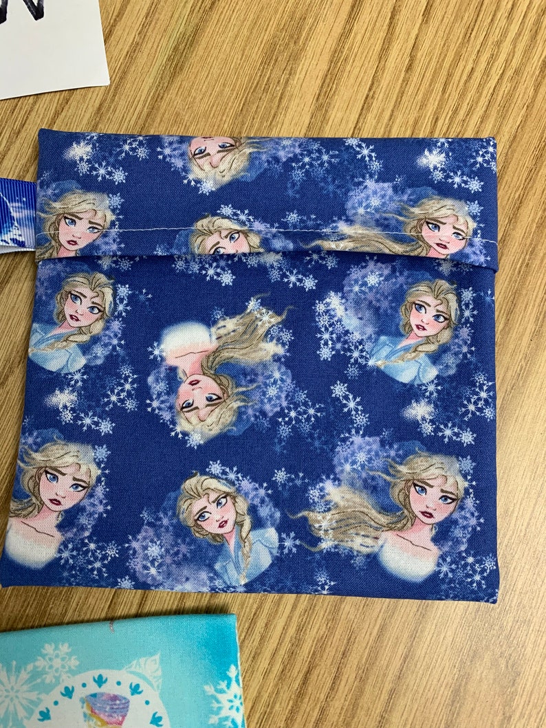 Op de afbeelding: Een blauwe stoffen tas met een wit sneeuwvlokkenpatroon en afbeeldingen van een cartoonfiguur met blond haar en blauwe ogen. De tas heeft een wit trekkoord.