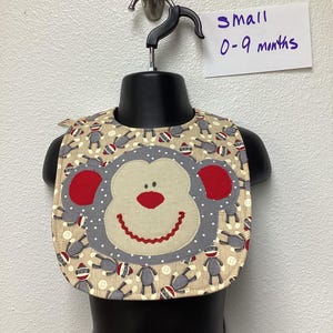 May include: A beige baby bib with a monkey face design. The monkey face is gray with red accents and a smiling mouth. The bib is decorated with small sock monkey figures. The tag reads "SMALL 0-9 months".