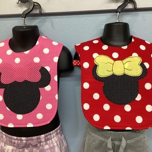 May include: Two baby bibs displayed on black mannequins. The left bib is pink with white polka dots and a black mouse silhouette with a pink bow. The right bib is red with white polka dots and a black mouse silhouette with a yellow bow.