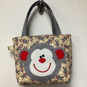 May include: A handmade tote bag with a monkey face applique. The bag features a tan background with a repeating pattern of sock monkeys. The monkey face is gray, white, and red, with black button eyes and a stitched smile. Gray handles complete the design.