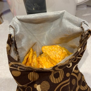 Op de afbeelding: Een bruine stoffen snacktas met een witte voering en een patroon van beige cirkels en strepen. De tas is gevuld met knapperige, oranje chips.