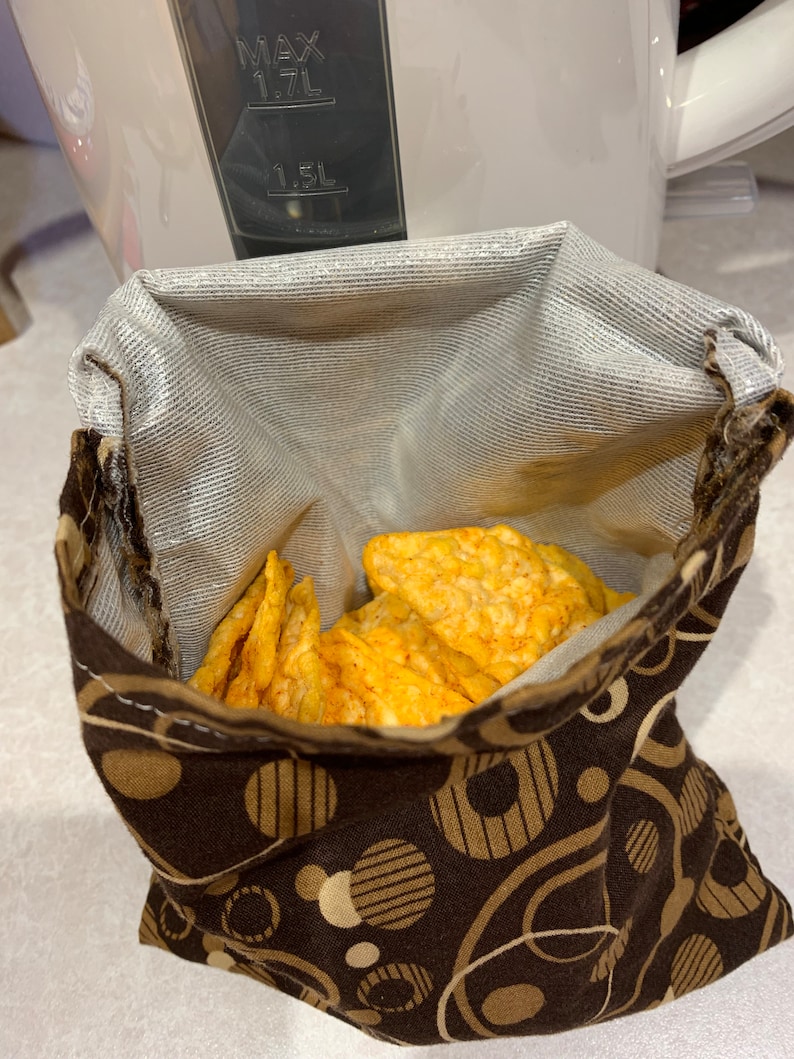 Op de afbeelding: Een bruine stoffen snacktas met een witte voering en een patroon van beige cirkels en strepen. De tas is gevuld met knapperige, oranje chips.