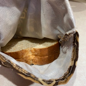 Op de afbeelding: Een bruine en beige stoffen tas met een witte voering. De tas is open en er liggen twee sneetjes brood in.