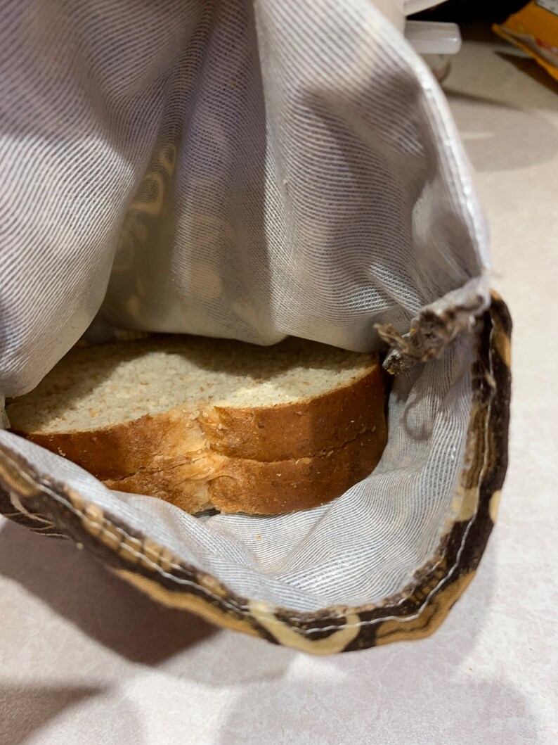 Op de afbeelding: Een bruine en beige stoffen tas met een witte voering. De tas is open en er liggen twee sneetjes brood in.