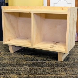 May include: A simple, unfinished wooden shelf unit with two open compartments and a top shelf. The shelf unit has four legs and is made of light-colored wood.