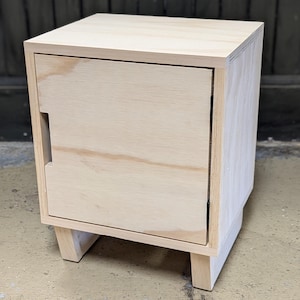 May include: A small, unfinished wooden nightstand with a single door and a simple design. The nightstand has a light wood finish and is made of plywood.