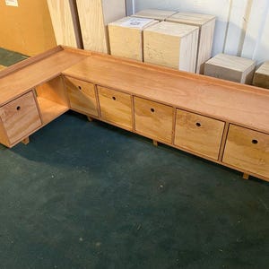 May include: A large, L-shaped wooden cabinet with a natural finish. It features a series of drawers and a top surface, ideal for storage or display. The cabinet is constructed from light-colored wood and has a simple, functional design.