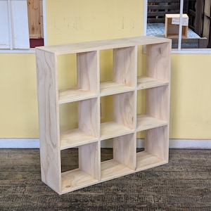 May include: A light-colored wooden shelving unit featuring a grid of nine square storage compartments arranged in three rows and three columns. The unit is constructed from unfinished light-toned wood with visible natural grain patterns throughout.