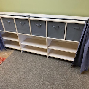 May include: A white wooden storage unit with six open shelves and six gray fabric storage bins. A black metal pipe rod is mounted across the top, featuring blue fabric curtains hanging from metal rings on both the left and right sides.
