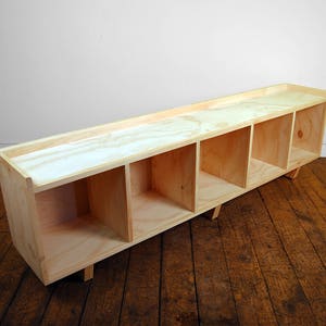 May include: A long, wooden shelf unit with four open compartments. The shelf is made of light-colored wood and has a simple, modern design.