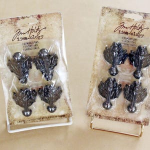 May include: Two packages of Jim Holtz Idea-ology Foundations, each containing four dark metal leaf-shaped decorative feet. The packaging has a vintage, distressed look with the word "Foundations" in English, French, and Spanish.