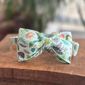 May include: A light blue bow tie with a repeating pattern of mushrooms, leaves, and berries. The bow tie is made of fabric and sits on a wooden surface. The design includes orange, green, and brown elements.