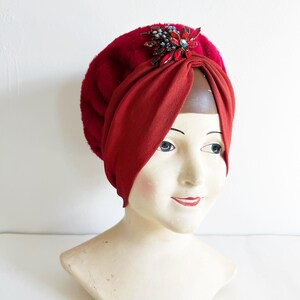 Peut inclure: Un turban bordeaux orné d'une broche décorative. Le turban présente une texture pelucheuse, semblable à de la fourrure, sur le dessus et un tissu lisse et drapé sur la partie inférieure. La broche est ornée de pierres rouges et turquoise.