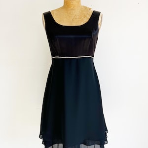 May include: A black cocktail dress with a velvet bodice and a flowing chiffon skirt. The dress features a delicate rhinestone trim at the waist and a scoop neckline. The dress is displayed on a vintage dress form.