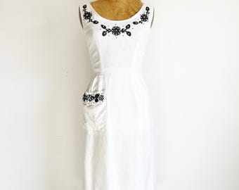 May include: A vintage white dress with black floral beadwork around the neckline and on the pocket. The sleeveless dress is displayed on a mannequin. The dress has a scoop neckline and a fitted bodice, with a knee-length skirt.