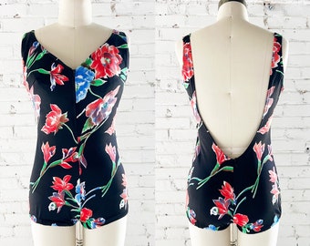 May include: A black one-piece swimsuit with a floral print featuring red, blue, and white flowers. The swimsuit has a low back and a V-neck.