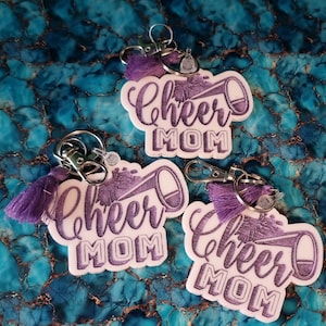 May include: Three white keychains with purple tassels and a purple glitter design. Each keychain features a white megaphone with the text "Cheer Mom" in purple glitter.