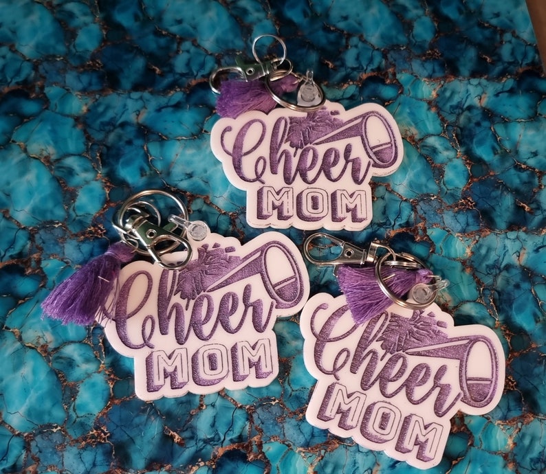 May include: Three white keychains with purple tassels and a purple glitter design. Each keychain features a white megaphone with the text "Cheer Mom" in purple glitter.
