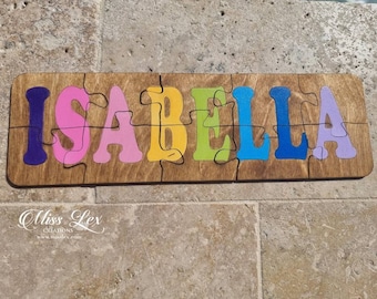 May include: A wooden puzzle with the name "ISABELLA" spelled out in colorful letters. Each letter is a separate piece of the puzzle. The letters are painted in different colors, including purple, pink, yellow, green, blue, and light purple.