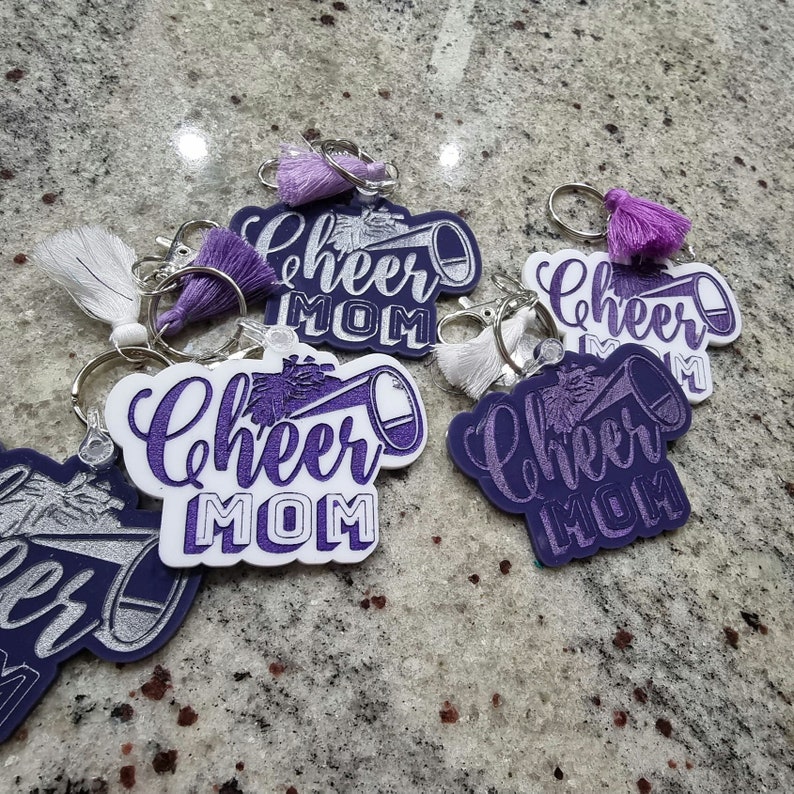 May include: Five purple and white keychains with the text "Cheer Mom" and a silver glitter pom-pom and megaphone design. The keychains have silver key rings and purple tassels.