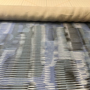 May include: A close-up of a blue and white patterned fabric with a subtle wave design. The fabric is rolled up and has a light brown color on the underside.