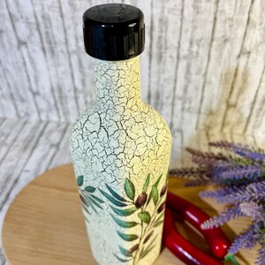 Puede incluir: Una botella de cer&aacute;mica blanca con una tapa negra y un dise&ntilde;o de rama de olivo verde. La botella est&aacute; sobre una superficie de madera.