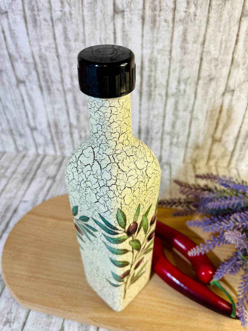 Puede incluir: Una botella de cer&aacute;mica blanca con una tapa negra y un dise&ntilde;o de rama de olivo verde. La botella est&aacute; sobre una superficie de madera.