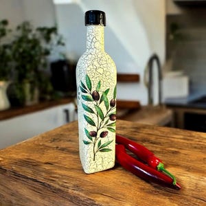 Puede incluir: Una botella de cer&aacute;mica blanca con tapa negra y un dise&ntilde;o de rama de olivo verde. La botella est&aacute; sobre una superficie de madera con dos chiles rojos.