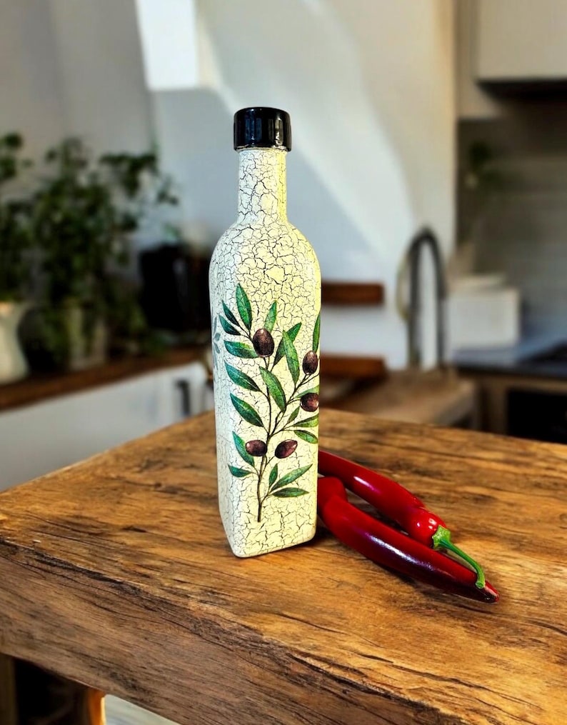 Puede incluir: Una botella de cer&aacute;mica blanca con tapa negra y un dise&ntilde;o de rama de olivo verde. La botella est&aacute; sobre una superficie de madera con dos chiles rojos.
