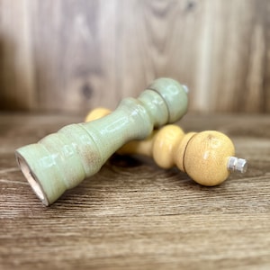 May include: Two wooden salt and pepper shakers, one is light green and the other is light yellow. The shakers are lying on a wooden surface.