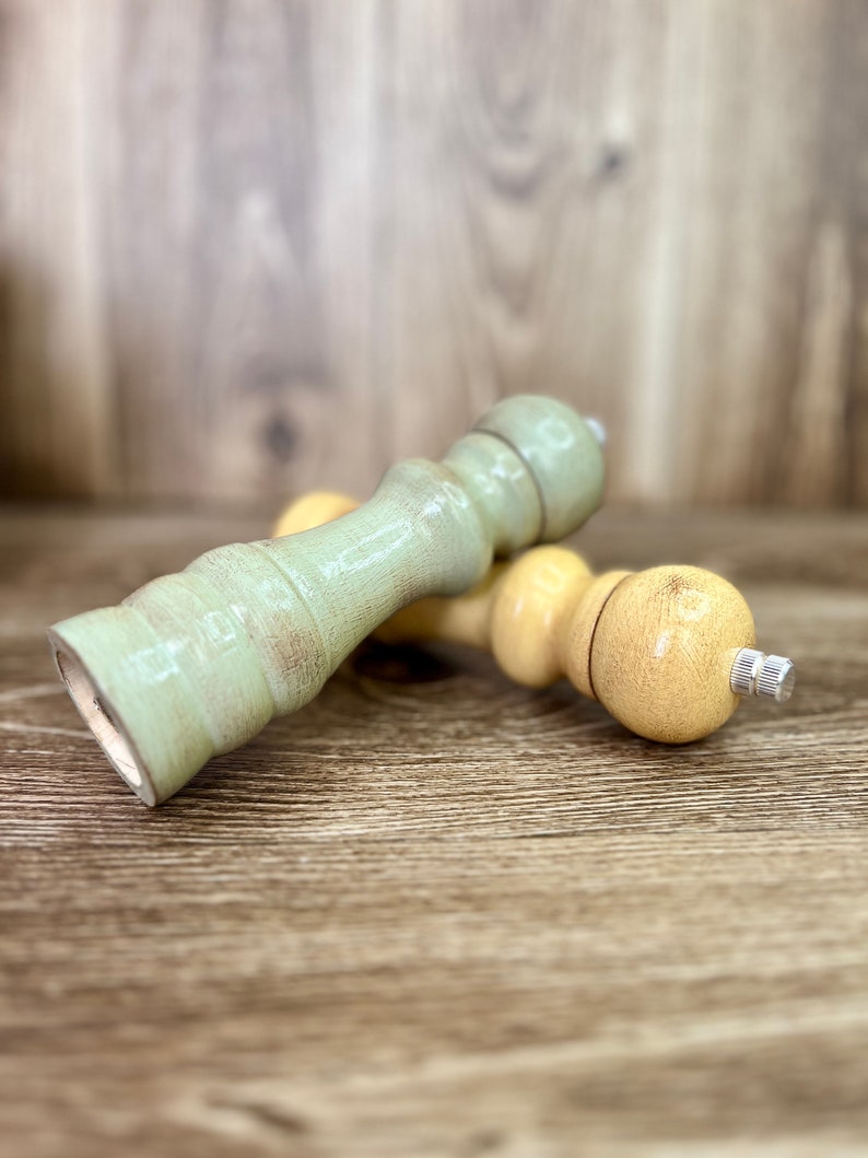 May include: Two wooden salt and pepper shakers, one is light green and the other is light yellow. The shakers are lying on a wooden surface.