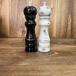 May include: Two wooden pepper and salt mills with a distressed finish. One mill is painted black with white worn edges, and the other is painted white with black worn edges. Both feature a rounded top, a curved body, and a silver metal knob.