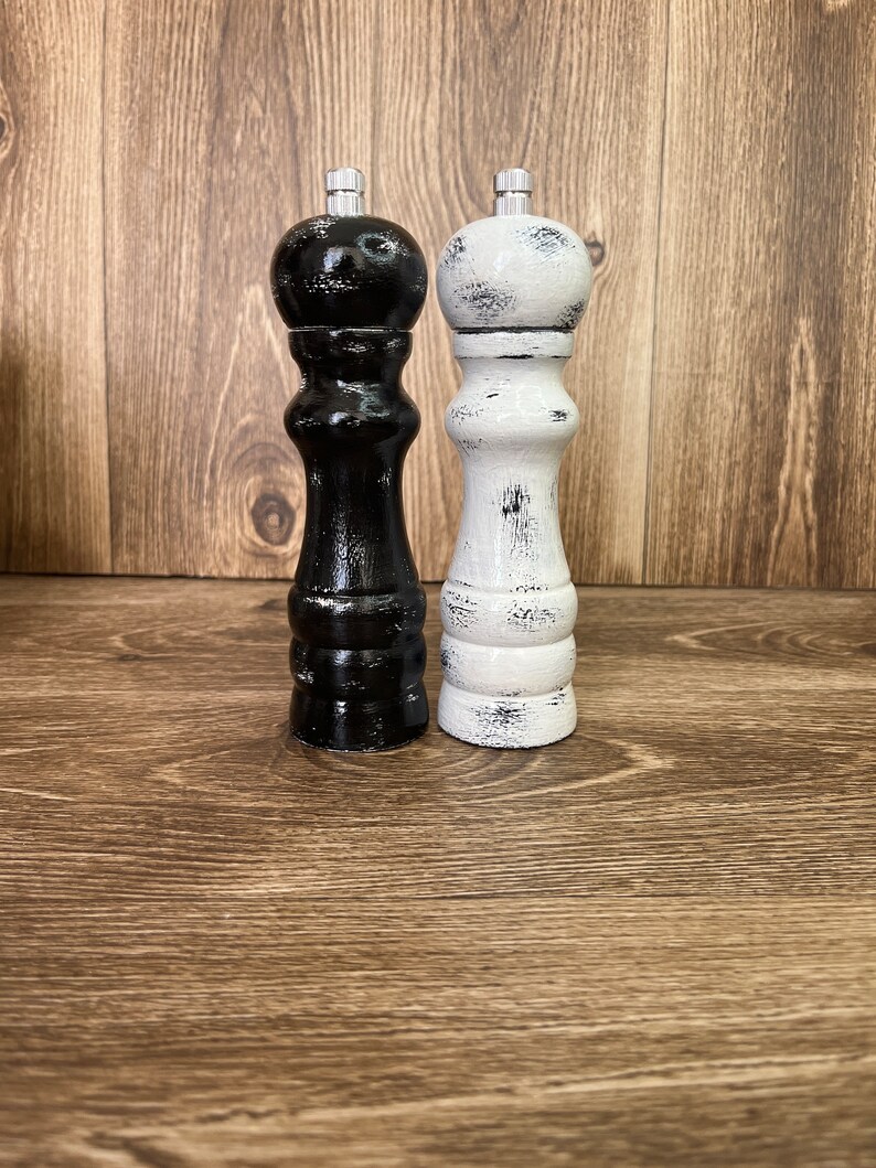 May include: Two wooden pepper and salt mills with a distressed finish. One mill is painted black with white worn edges, and the other is painted white with black worn edges. Both feature a rounded top, a curved body, and a silver metal knob.