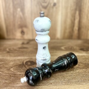 May include: A pair of wooden pepper mills with a distressed finish. One mill is painted white with black wear marks, and the other is painted black with white wear marks. Both feature a silver-toned metal knob at the top for grinding.