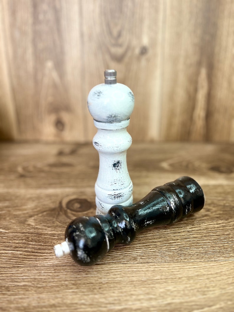 May include: A pair of wooden pepper mills with a distressed finish. One mill is painted white with black wear marks, and the other is painted black with white wear marks. Both feature a silver-toned metal knob at the top for grinding.
