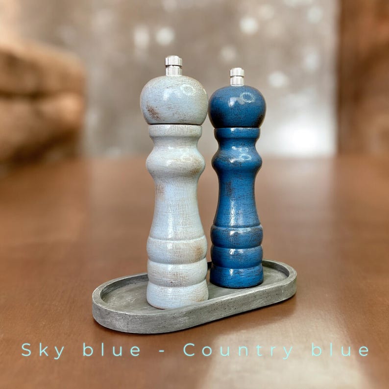 May include: Two salt and pepper shakers on a gray tray. The shaker on the left is painted a light blue color and the shaker on the right is painted a darker blue color. The text "Sky blue - Country blue" is printed below the shakers.