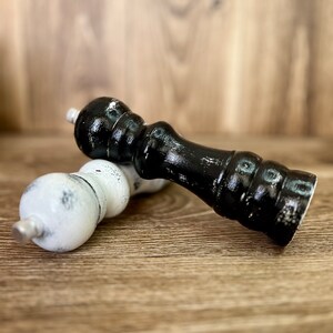 May include: Two wooden pepper mills, one finished in glossy black and the other in white, both featuring a distressed paint effect. Each mill has a rounded top, a contoured body with horizontal ridges, and a small metallic knob at the apex.