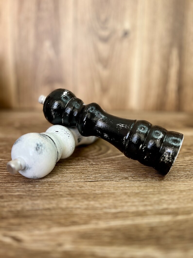 May include: Two wooden pepper mills, one finished in glossy black and the other in white, both featuring a distressed paint effect. Each mill has a rounded top, a contoured body with horizontal ridges, and a small metallic knob at the apex.
