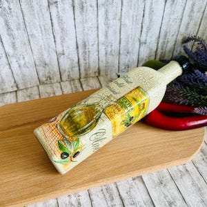 Può includere: Una bottiglia di olio d'oliva decorativa con una finitura bianca invecchiata e un'etichetta che recita "First cold pressed Extra Virgin Olive Oil". La bottiglia è appoggiata su un tagliere di legno.