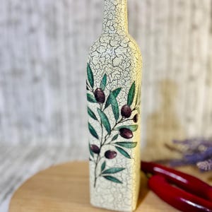 Puede incluir: Una botella de cer&aacute;mica blanca con tapa negra y un dise&ntilde;o de ramas de olivo con hojas verdes y aceitunas moradas. La botella est&aacute; sobre una superficie de madera.