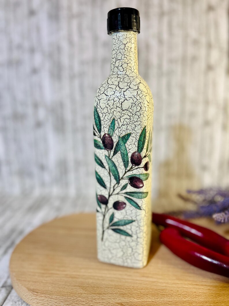 Puede incluir: Una botella de cer&aacute;mica blanca con tapa negra y un dise&ntilde;o de ramas de olivo con hojas verdes y aceitunas moradas. La botella est&aacute; sobre una superficie de madera.