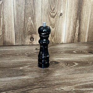 May include: A tall, black pepper mill with a glossy finish and a cylindrical, hourglass-shaped body. The top features a small, metallic silver adjustment knob. The design is smooth and uniform, standing upright with a rounded top section.