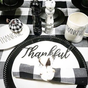 May include: A table setting featuring a white plate with the word Thankful in black script, a black and white buffalo check napkin with a cotton boll napkin ring, a white mug with Thankful text, and a small buffalo check pumpkin.