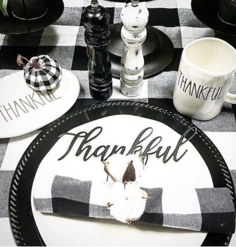 May include: A table setting featuring a white plate with the word Thankful in black script, a black and white buffalo check napkin with a cotton boll napkin ring, a white mug with Thankful text, and a small buffalo check pumpkin.