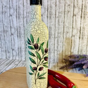 Puede incluir: Una botella de vidrio decorativa con tapa negra y acabado blanco craquelado. La botella presenta un dise&ntilde;o de rama de olivo verde con aceitunas negras.