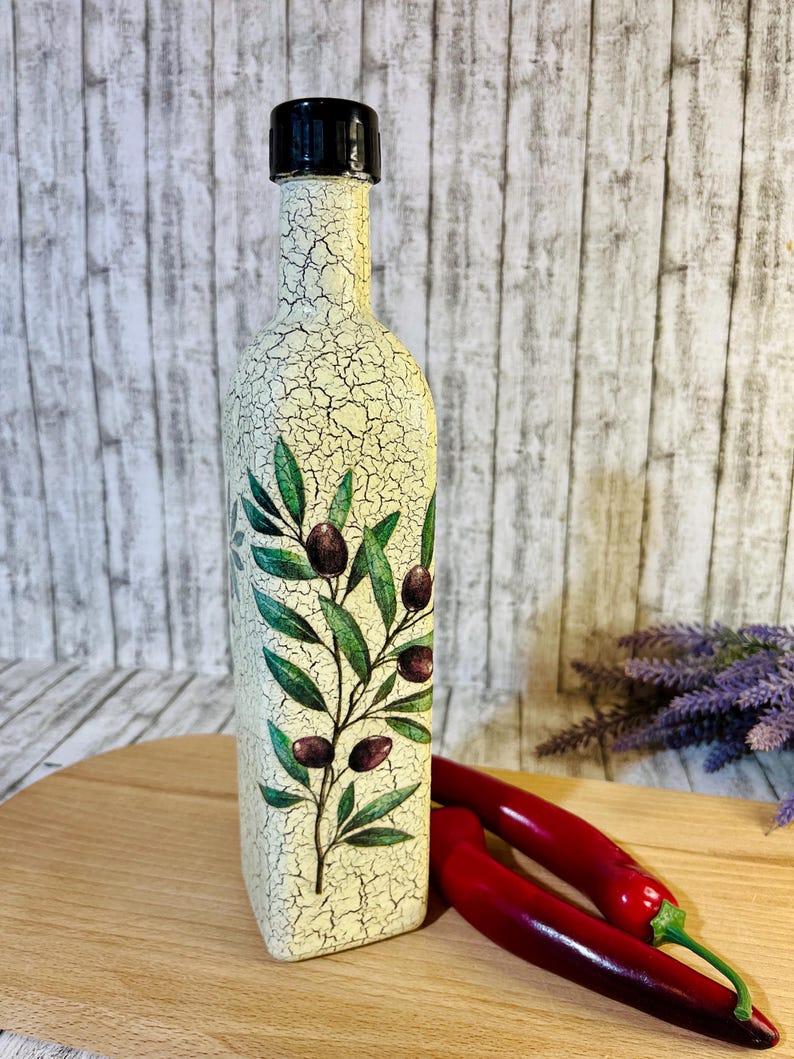 Puede incluir: Una botella de vidrio decorativa con tapa negra y acabado blanco craquelado. La botella presenta un dise&ntilde;o de rama de olivo verde con aceitunas negras.