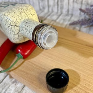 May include: A white ceramic bottle with a crackled finish and a black and clear plastic pour spout. The bottle is sitting on a wooden cutting board with two red chili peppers.